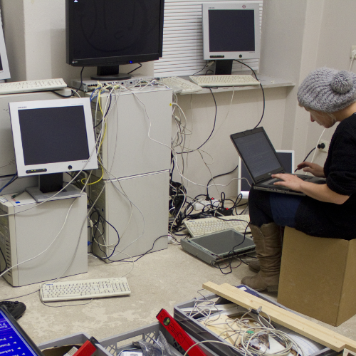 In einem chaotischen Raum mit Werkzeugen und vielen Computern arbeitet eine junge Frau an Quellcode.