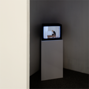 Auf einem Sockel steht ein Fernseher in Kubus-Form. Im Videobild steht ein Holztisch, darauf sitzend lehnt eine Frau ihren Kopf an einen großen weißen Würfel.