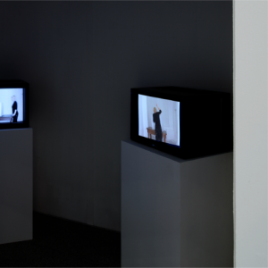 2 Fernseher in Kubus-Form stehen auf weißen Sockeln. Nur der rechte Fernseher ist komplett im Bild: Im Video erhebt eine Frau mit schwarzer Kleidung den Arm.