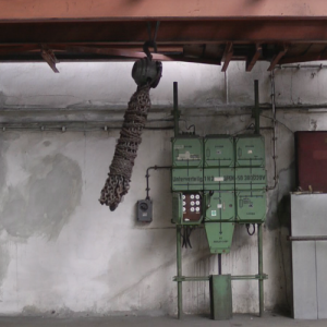 In einer Werkshalle mit alten Strom-Verteilerkästen schwingt ein riesiger Strang rostiger Ketten nach links.