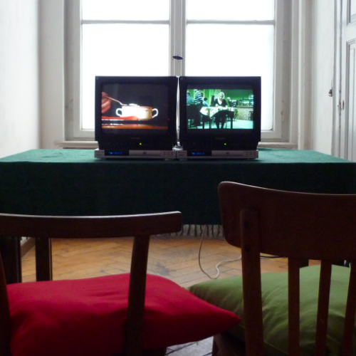Auf einem Tisch stehen 2 Fernseher eng nebeneinander. Die Video-Bilder passen farblich zu den Kissen der 2 Stühle vor dem Tisch: links rot, rechts grün.