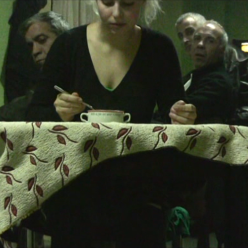 Eine junge Frau sitzt in einem Raum mit grünlichem Licht und löffelt aus einer Suppenschale. 3 Männer sehen ihr konzentriert dabei zu.