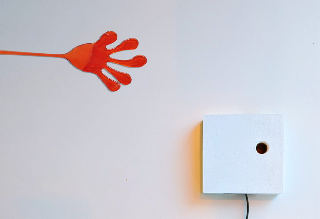 Eine Spielzeughand aus glibberigem, orangefarbenem Silikon haftet quer an einer Wand. Daneben hängt ein Kästchen mit einem nach unten führenden Kabel.