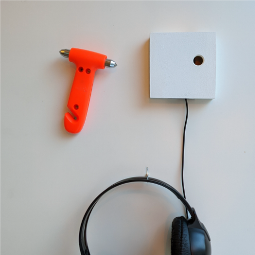 Neben einem orangefarbenen Nothammer hängen ein Vollkopfhörer und ein kleiner weißer Kasten an der Wand.