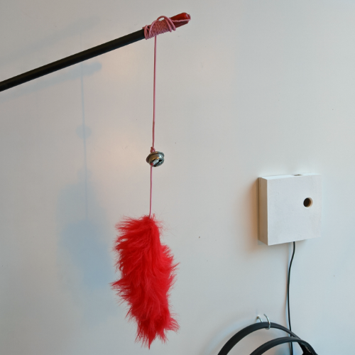 Eine Katzen-Angel mit Glöckchen und rotem Plüsch-Wurm am Faden, daneben hängt ein quadratisches Kästchen mit einem Kabel.