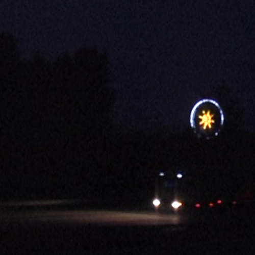 Nur die Scheinwerfer von einem LKW und ein riesiger Stern in einem Kreis über dem Fahrzeug leuchten in dieser Nachtaufnahme. Schemenhaft sind die Fahrspur und Bäume am Rand zu erkennen.