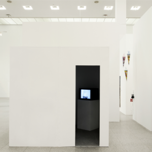 In einer Ausstellungshalle steht ein makelloser weißer Würfel mit schmalem Eingang als Raum im Raum. Aus dem dunklen Inneren leuchtet ein Video.
