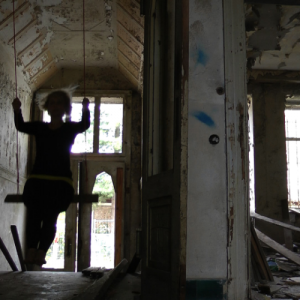 Im Treppenbereich von einem stark verfallenen Wohnhaus liegen Trümmer und Müll herum. Eine Frau, nur als schwarzer Schemen erkennbar, schaukelt mit großem Schwung durch den Raum.
