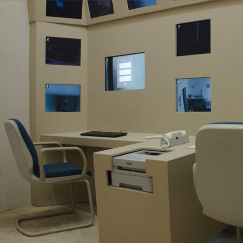 Ein Arbeitsplatz mit einer Tastatur und 9 in einer Wand eingelassenen Monitoren. In der Mittelkonsole zum benachbarten Arbeitsplatz sind Drucker und Telefon eingebaut.