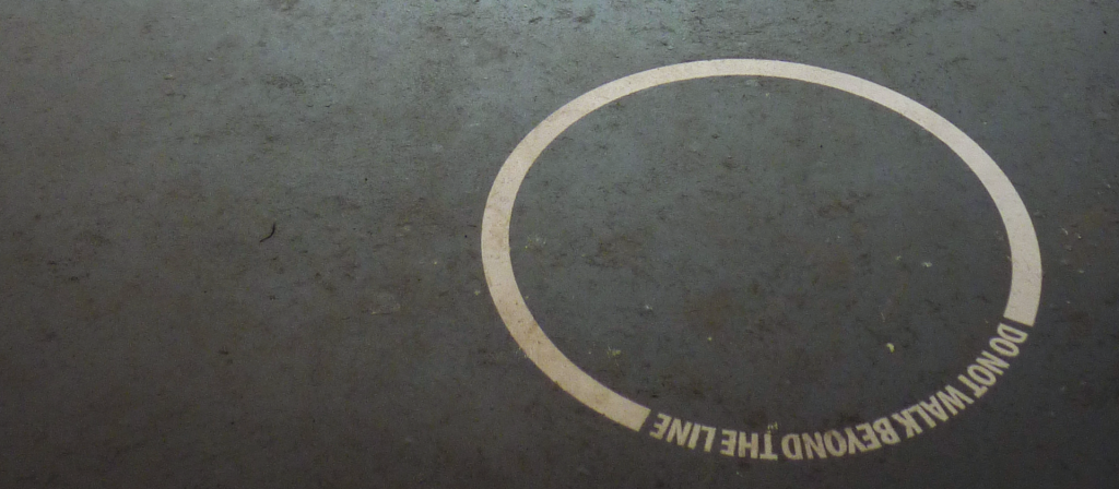 Auf dunkelgrauem Asphalt ist eine weiße ringförmige Markierung aufgebracht. Die Worte "Do not walk beyond the line" in Din-Schrift bilden zusammen mit einer breiten Linie einen geschlossenen Kreis.