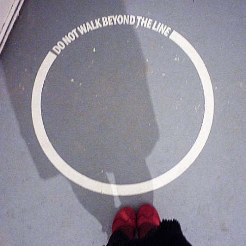 Der Schatten einer Person mit roten Schuhen fällt auf eine ringförmige Boden-Markierung mit der Inschrift: Do not walk beyond the line.