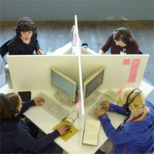 Ein quadratischer Tisch wird von diagonal aufgestellten Trennwänden in einzelne Arbeitsplatz-Nischen unterteilt. 4 junge Frauen mit Headsets arbeiten konzentriert an ihren Computern.