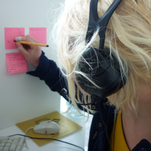 Eine blonde, junge Frau mit Headset notiert etwas Unleserliches auf einen von drei pinken Zetteln, die an einer weißen Trennwand direkt an ihrem Arbeitsplatz kleben.