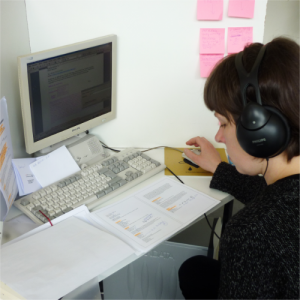 Eine Frau mit kurzen, dunklen Haaren liest in ausgedruckten, handschriftlich kommentierten Dokumenten, während auf ihrem Monitor ein Formular abgespeichert wird.