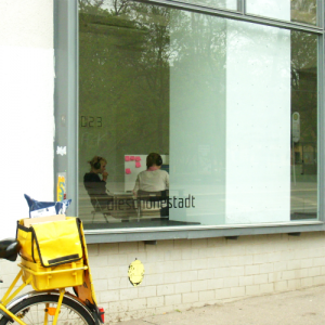Ein Post-Fahrrad steht vor dem Schaufenster der Galerie „Die schöne Stadt“. Im Galerie-Raum sitzen 2 Personen mit Headsets an einem Tisch. An der Wand, die die beiden Arbeitsplätze trennt, kleben pinke Haftnotizen.