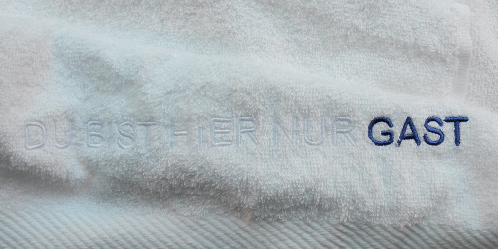 Zweifarbig besticktes Handtuch. Im weißen Farbton des Handtuchs erscheinen die ersten Worte der Stickerei: „Du bist hier nur“. „Gast“, das letzte Wort, kontrastiert in dunkelblau.