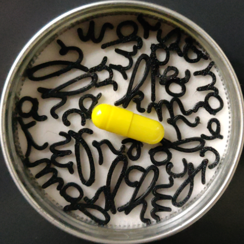 In einer silbernen Tablettendose liegt mittig eine knallgelbe Kapsel, um sie herum ein Durcheinander aus schwarzen kursiven Kleinbuchstaben.