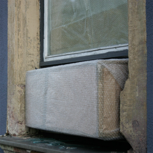 Ein Quader aus Luftpolsterfolie presst sich aus einem Fenster, auf dem Fensterbrett liegt herausgebrochenes Glas.