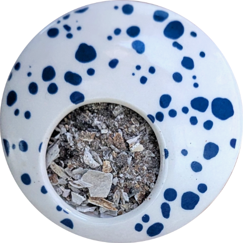 Asche mit kleinen Knochen-Fragmenten darin liegt in einer weiß-blauen Porzellan-Form mit einer kreisrunden Öffnung jenseits der Mitte, fast wie ein Auge.